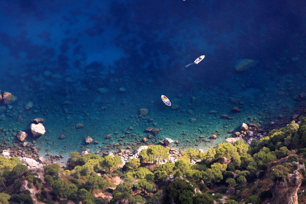 Aerial view of small boats on clear blue water beside a rocky, wooded coast